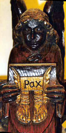 Angel holding a wooden shield on which are engraved the shoes of peace below the word Pax
