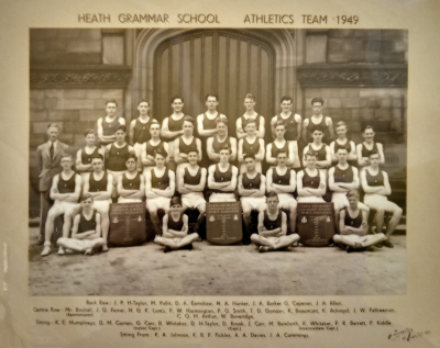 Formal photograph showing four rows of athletes, two rows standing with Mr Birchall (Sportsmaster) to the left of the forward row, one row seated and four athletes sitting crosslegged in front with three trophies spread out between them